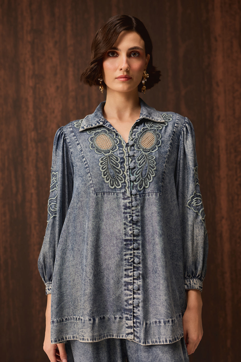 A model stands against a dark wood-paneled wall wearing a matching light-wash denim two-piece set consisting of a long-sleeve button-front top with decorative cutouts and embroidery, and wide-leg denim pants. She is wearing lime green pointed-toe heels.