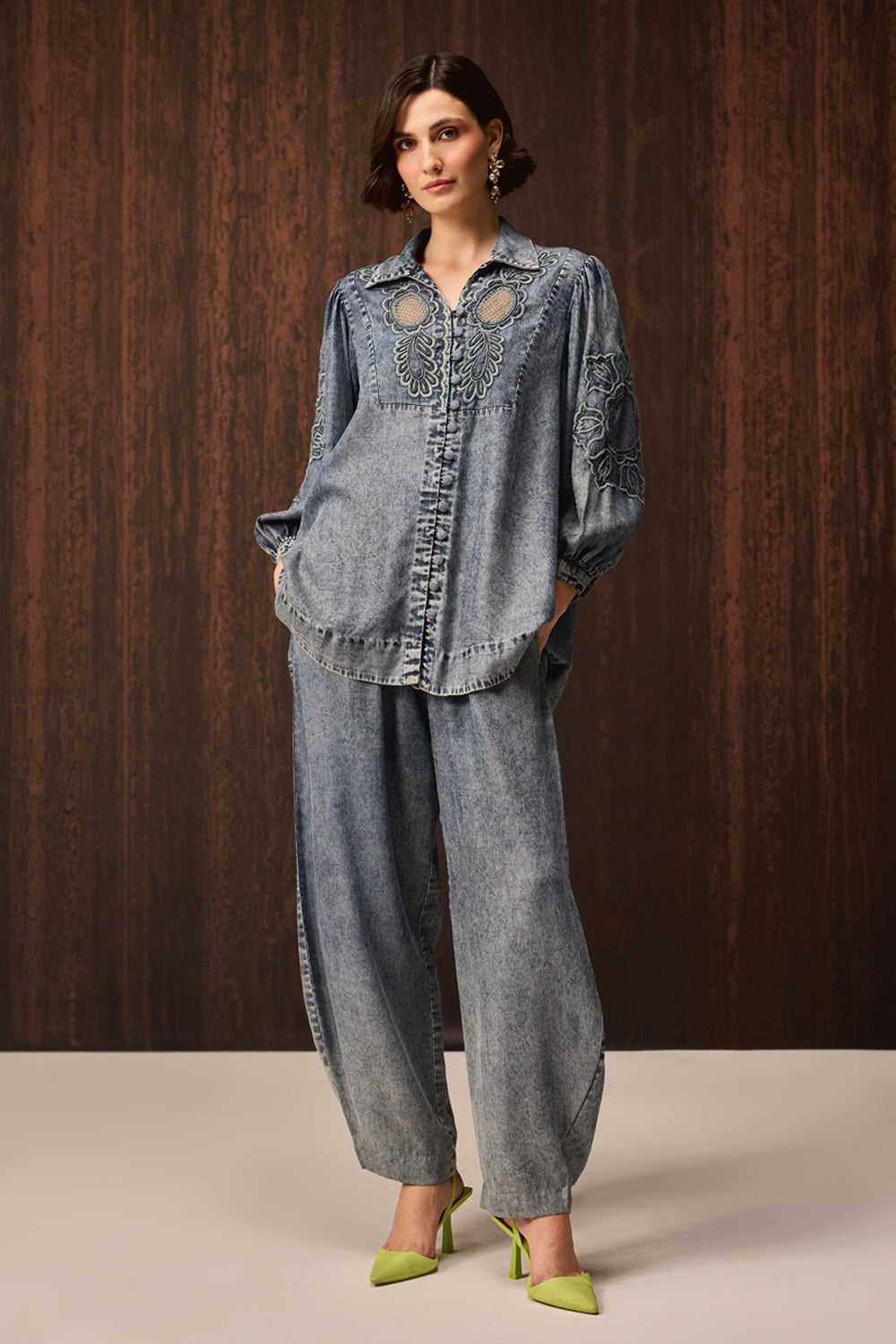 A model stands against a dark wood-paneled wall wearing a matching light-wash denim two-piece set consisting of a long-sleeve button-front top with decorative cutouts and embroidery, and wide-leg denim pants. She is wearing lime green pointed-toe heels.