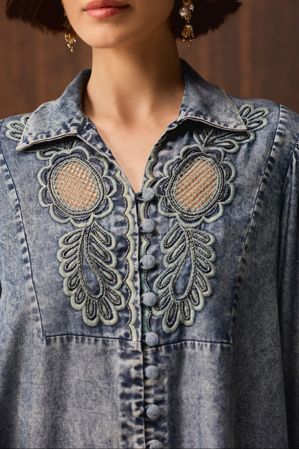 Close-up of a person wearing a blue denim button-front top with ornate embroidered detailing on the chest and collar, against a warm brown background.