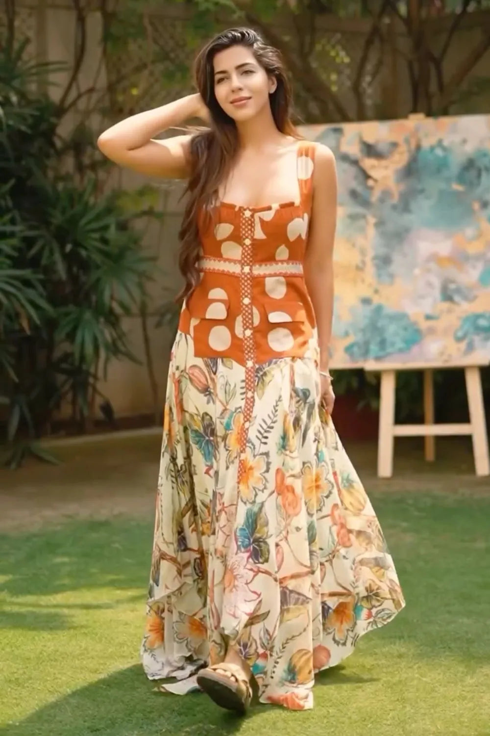 Woman in a patterned dress standing outdoors with plants and a colorful painting in the background