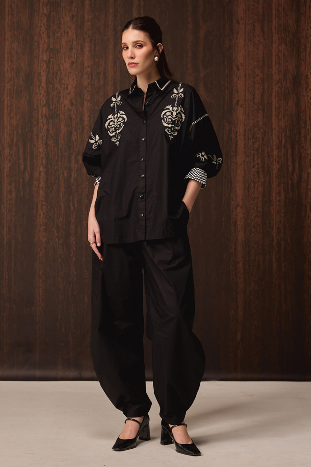 A model stands against a dark wood-paneled background wearing a black button-up shirt with ornate white embroidery on the chest and sleeves, paired with loose black pants and black heels.