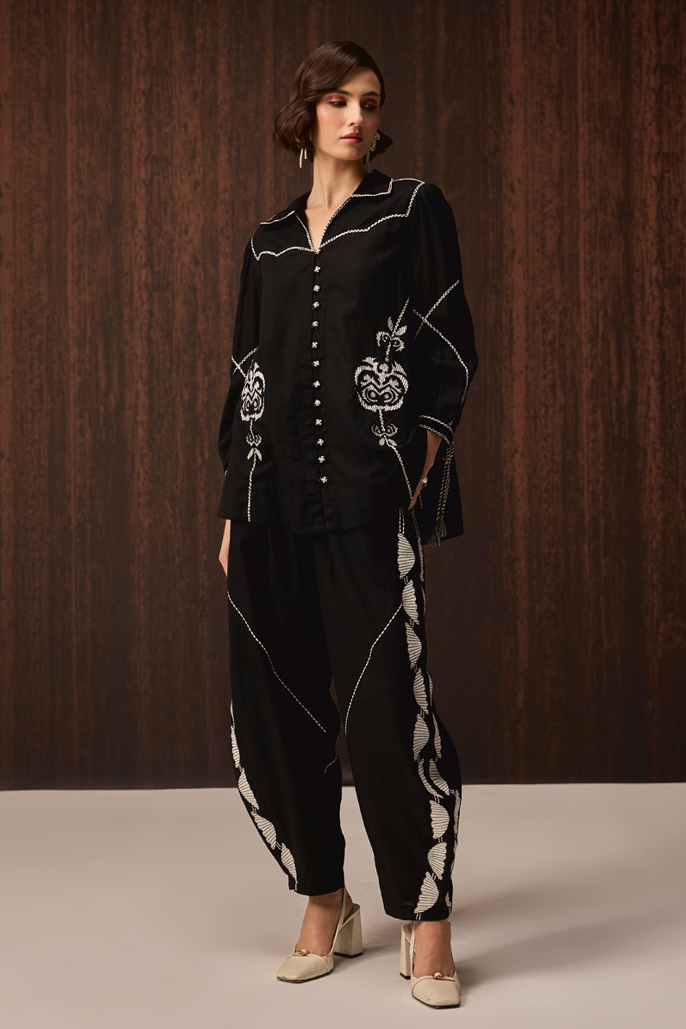 A woman models a black embroidered two-piece outfit consisting of a long-sleeve button-front top and matching loose pants, with white contrast stitching and embroidery details, set against a dark wood-paneled background.