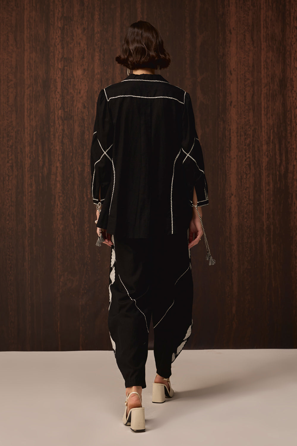 A model viewed from behind wearing a black two-piece outfit with white contrast stitching, consisting of a long-sleeve top and matching loose pants, walking against a dark wood-paneled background.