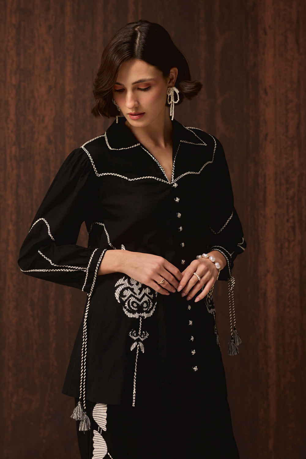 A woman wearing a black long-sleeve embroidered tunic top with white decorative stitching, buttons, and tassels, posed against a dark brown wood-paneled background.
