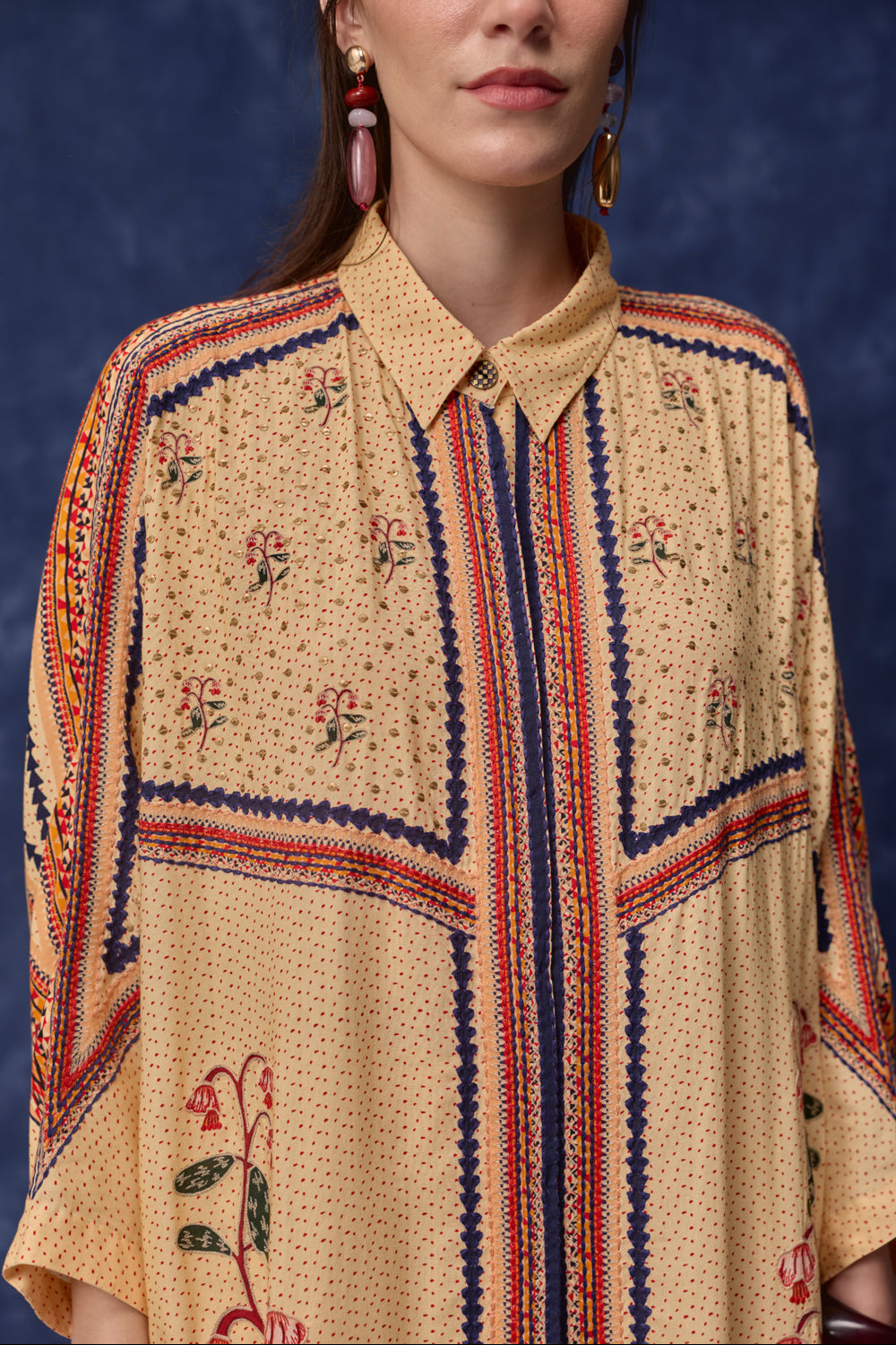 A close-up of a model wearing a beige button-up shirt with a dense micro-print, decorated with navy blue, red, and green geometric borders and floral motifs, set against a dark blue textured background.