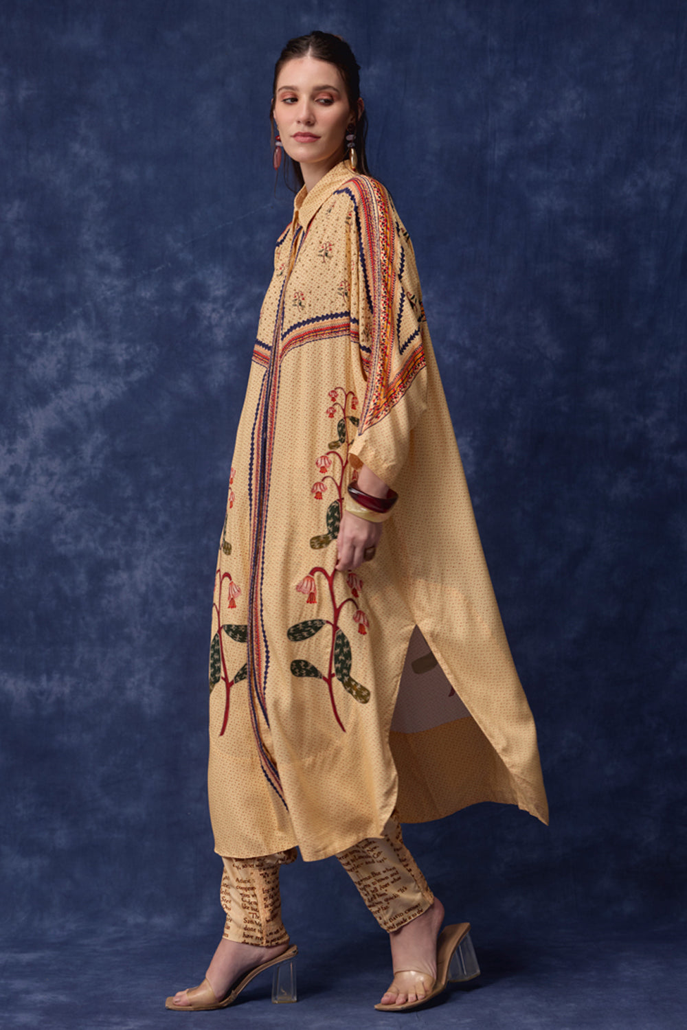 A woman models a beige embroidered kurta with floral patterns and matching patterned pants against a textured dark blue studio backdrop.