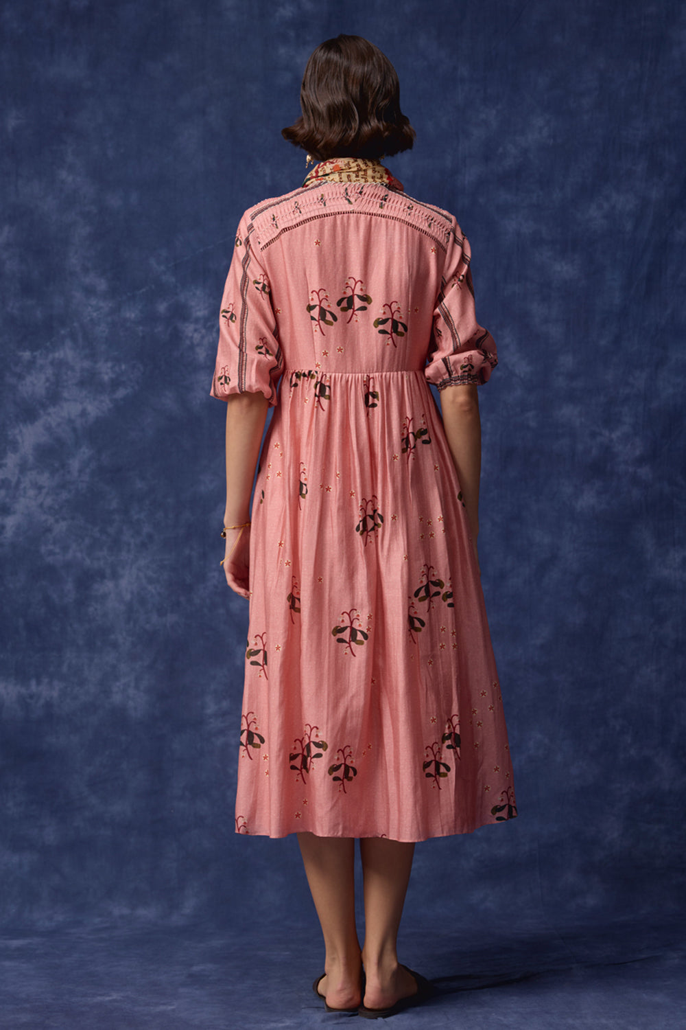 A woman stands with her back to the camera wearing a pink midi dress with black embroidered floral motifs and puff sleeves, set against a textured blue studio backdrop.
