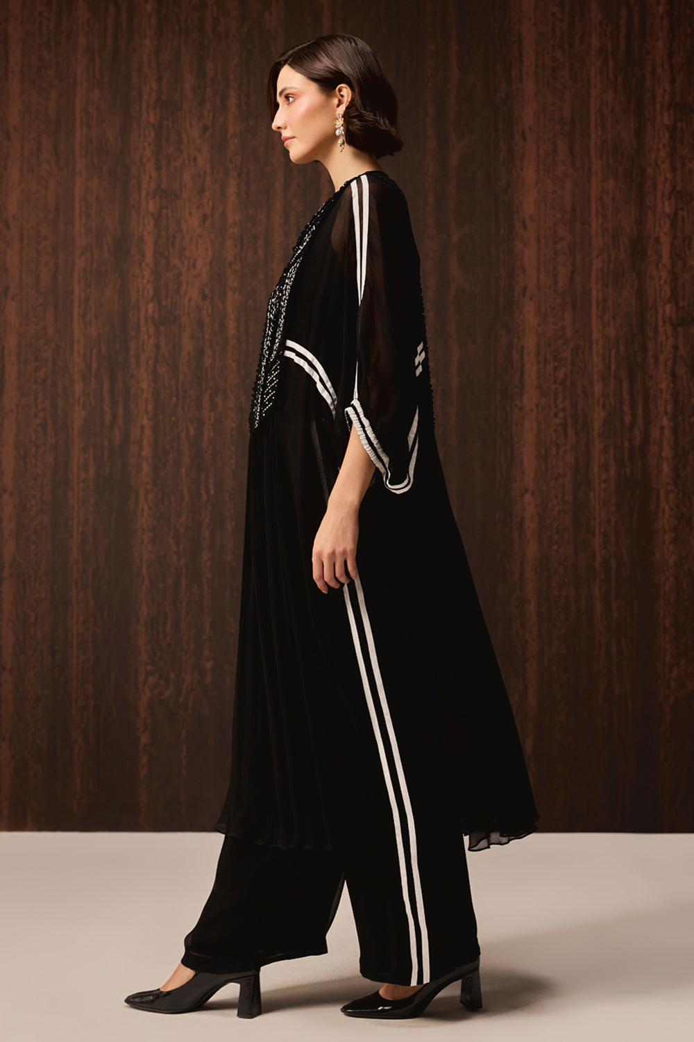 A woman models a black sheer tunic with white stripe details and matching black wide-leg trousers against a dark wood-paneled background.