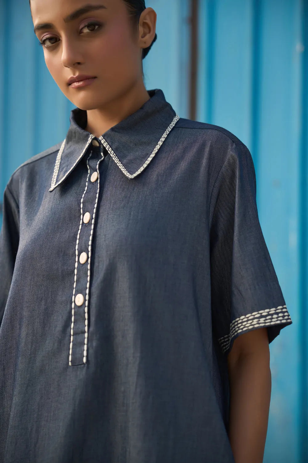 Closeup look of Rico Mini Dress women's dark blue chambray mini dress featuring a sharp pointed collar, short sleeves, a partial button-down front and contrast white stitching along the collar, sleeves, button placket and hem, finished with a loose flared A-line silhouette for casual outings, summer travel, relaxed daytime wear and casual dinners.