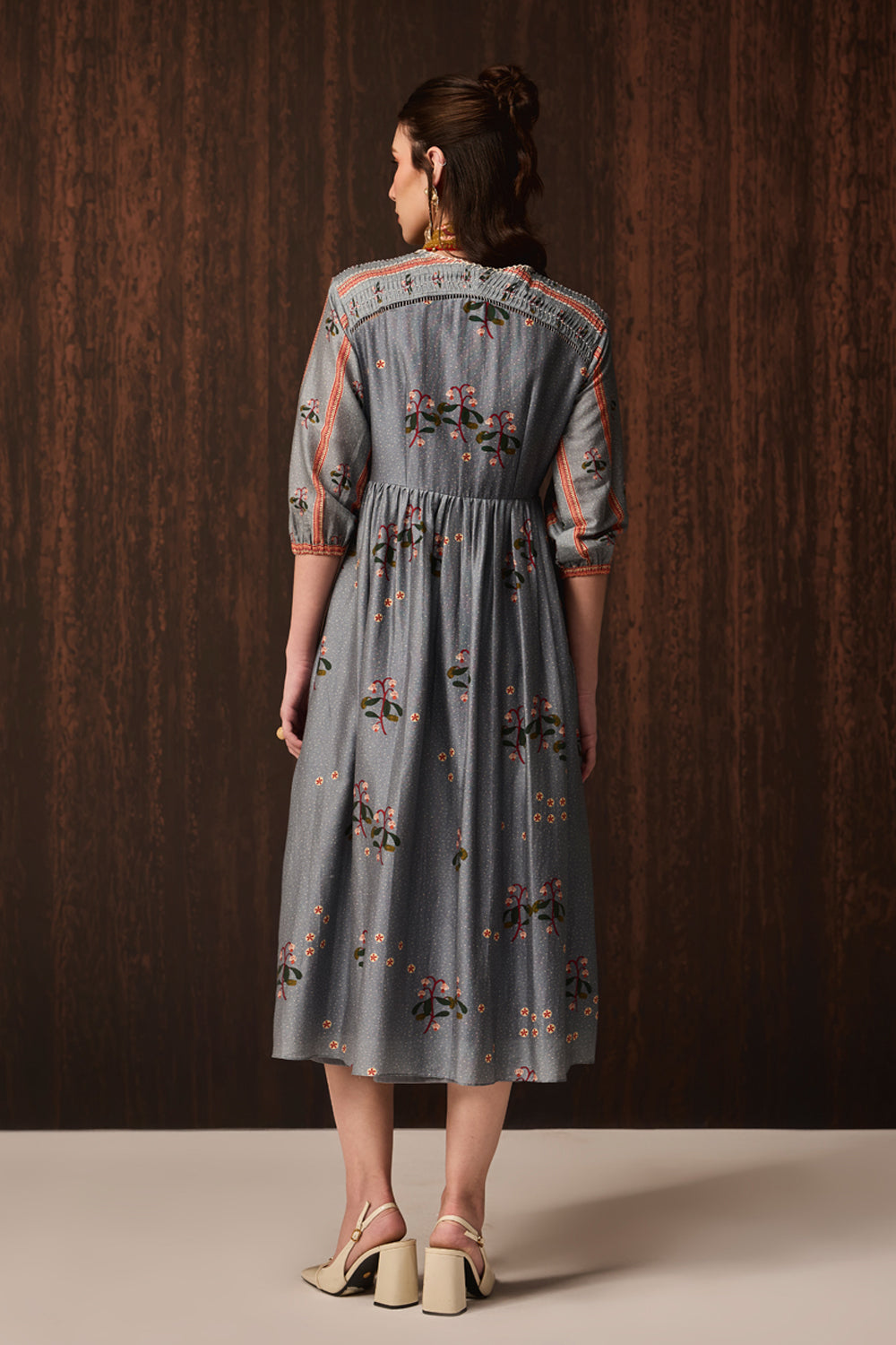 A woman viewed from the back wearing a gray midi dress with colorful floral embroidery and 3/4 sleeves, standing against a dark wood-paneled background.