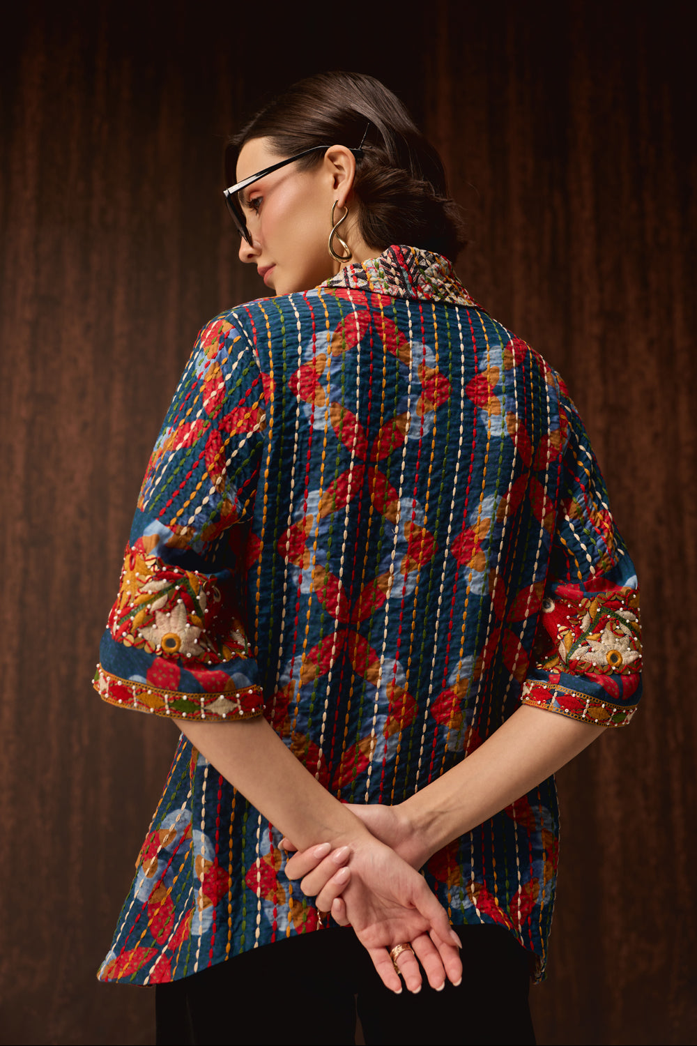A woman shown from the back wearing a colorful patterned shirt with a geometric design in red, blue, yellow, and white, paired with black pants against a dark wood background.