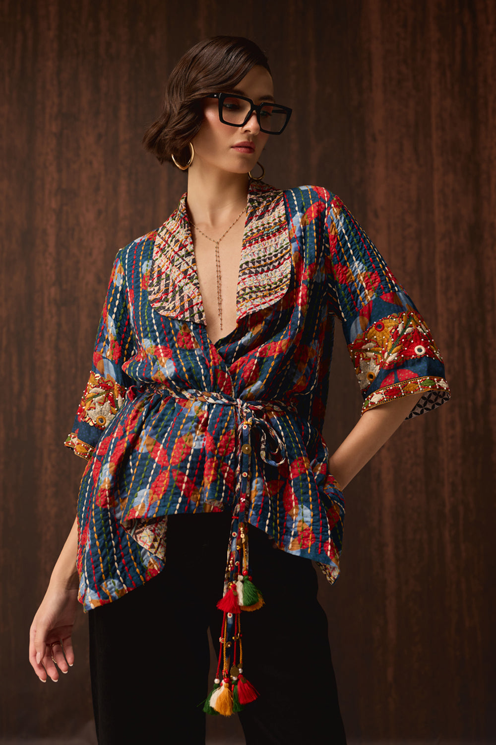 A woman wearing a colorful patterned wrap-style blouse with tassel ties and black pants, posed against a dark wood-paneled background.