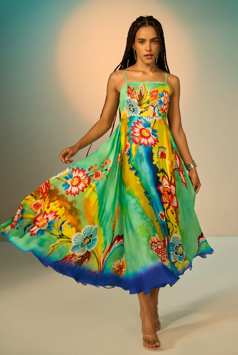 A woman modeling a vibrant, flowing midi dress with a colorful floral and abstract print. The dress features thin spaghetti straps and a ruffled hem, shown as she twirls against a studio backdrop with a warm spotlight.