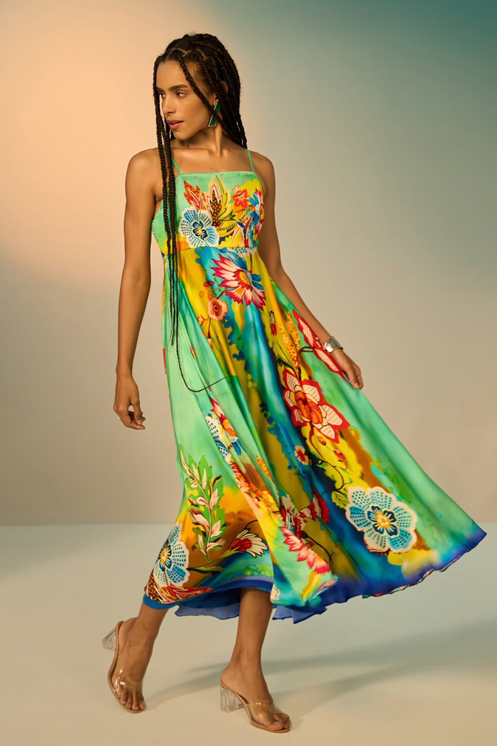 A woman modeling a sleeveless, flowy midi dress with a bright tropical floral print in shades of green, yellow, blue, orange, and pink against a soft gradient studio background.