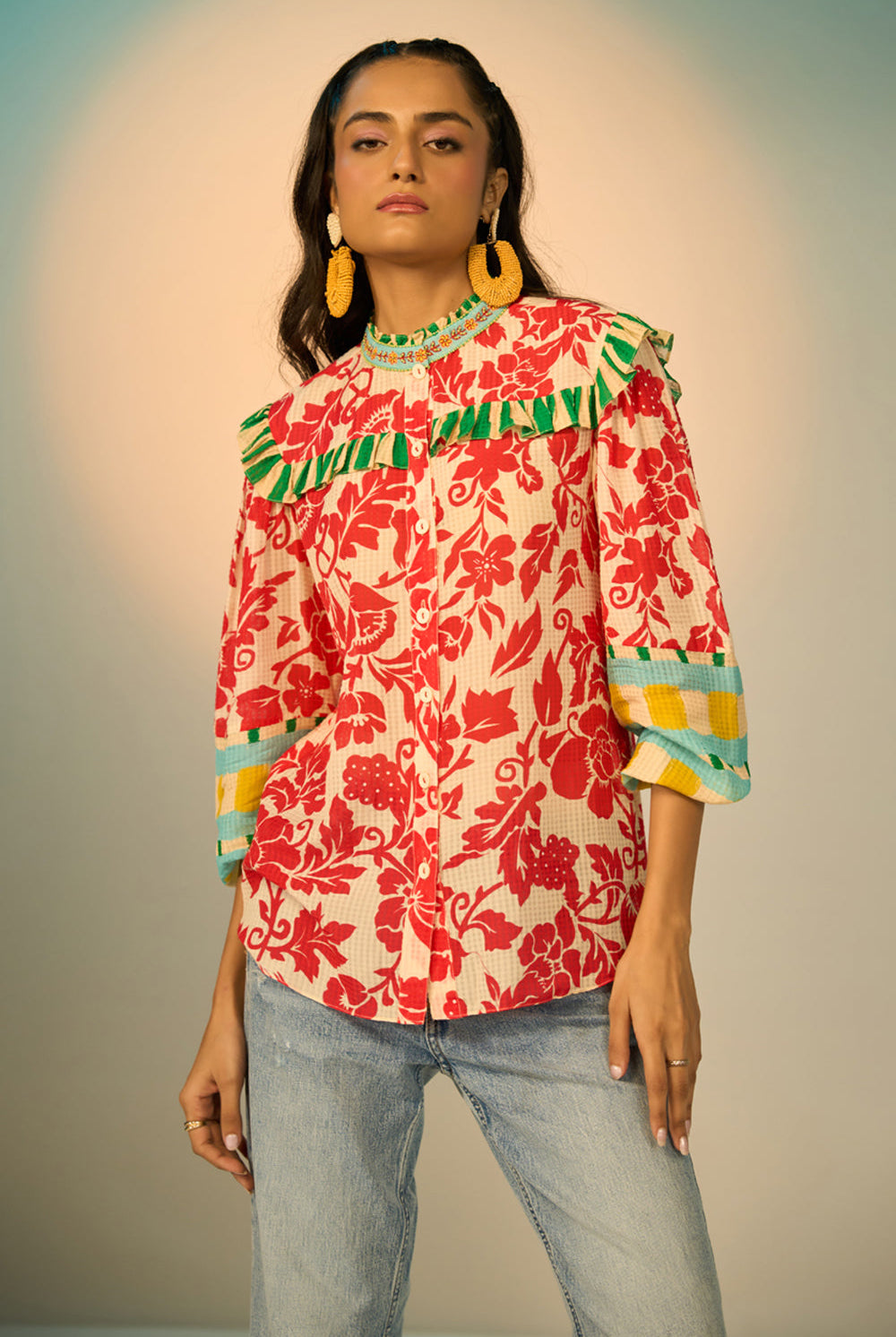 A woman modeling a vibrant red and cream floral print blouse with green and yellow accents, paired with light-wash jeans and gold hoop earrings, set against a soft beige studio background.