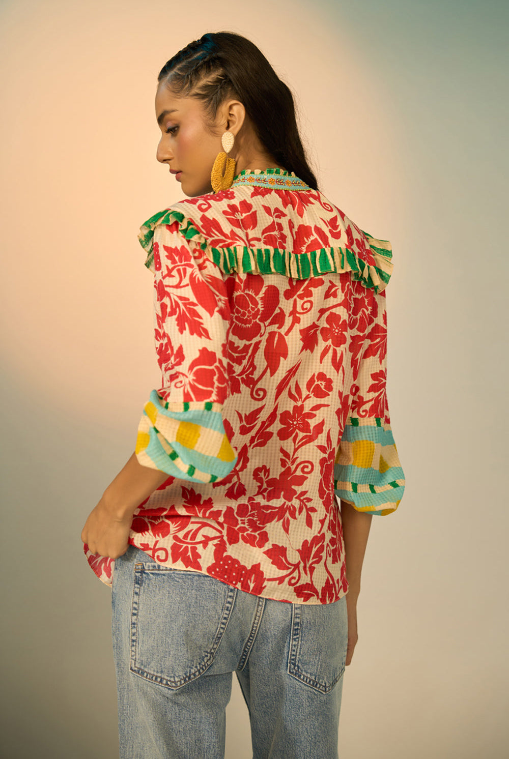 A woman modeling a vibrant red and cream floral print blouse with green and yellow accents, paired with light-wash jeans and gold hoop earrings, set against a soft beige studio background.