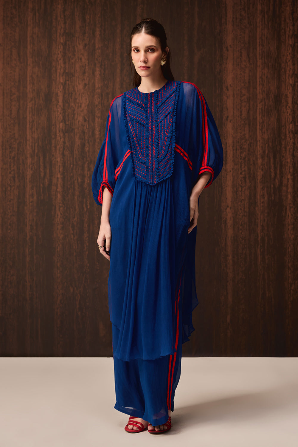 A woman models a royal blue tunic and trouser set with red and navy embroidered details on the chest and red piping along the sleeves and side seams, set against a dark wood-paneled background.