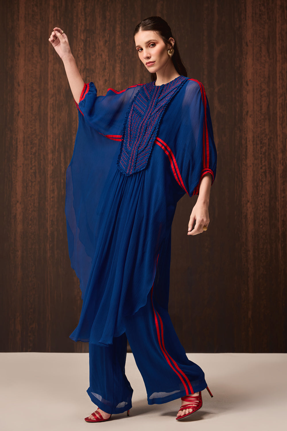 A woman models a royal blue draped tunic and trouser set with red and navy embroidered details, set against a dark wood-paneled background.