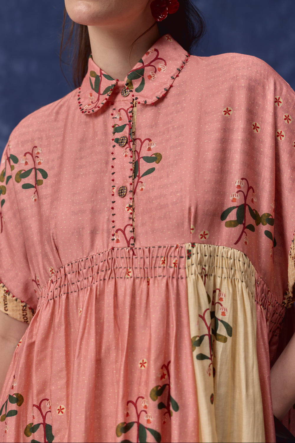 A close-up of a woman wearing a dusty pink cotton dress with floral embroidery, a Peter Pan collar, and a smocked waist seam. The dress features a cream-colored smocked panel on the skirt and is shown against a dark blue textured background.