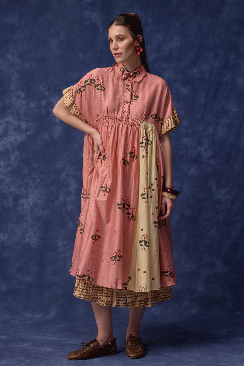 A woman models a dusty pink cotton dress with black floral embroidery, a beige side panel, and gold lace trim, standing against a textured blue studio backdrop.