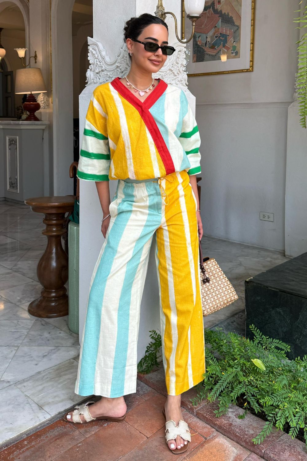 Arushi Mehra in a colorful striped outfit standing in an outdoor setting with plants and a white wall.