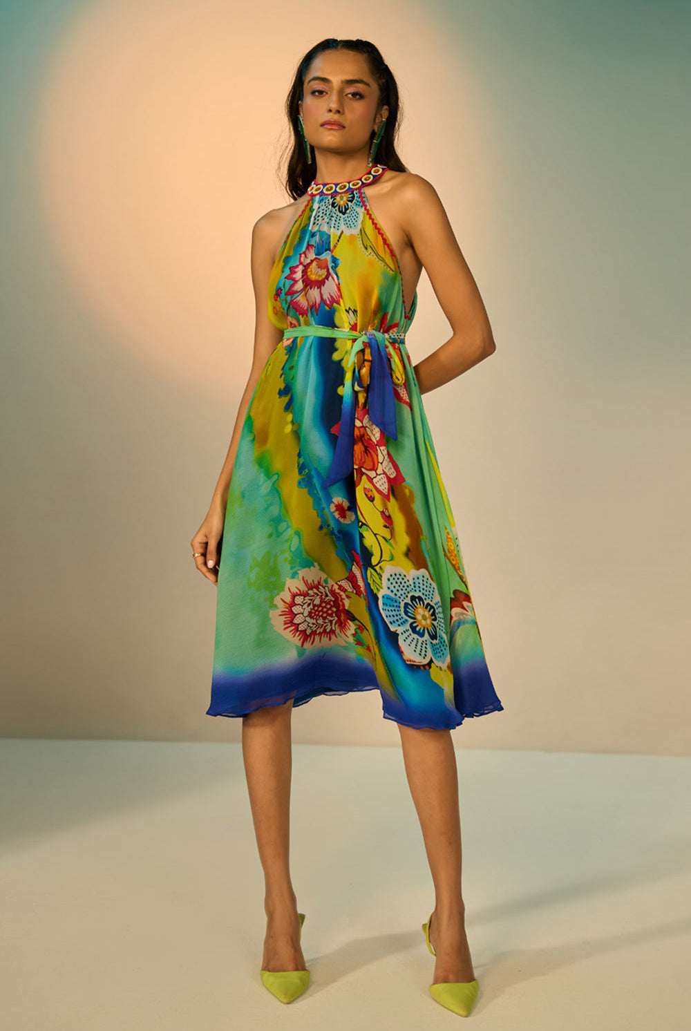 A woman modeling a sleeveless halter-neck midi dress with a vibrant multicolor floral and abstract print in shades of blue, green, yellow, and pink, styled with lime green pointed-toe heels against a studio backdrop.