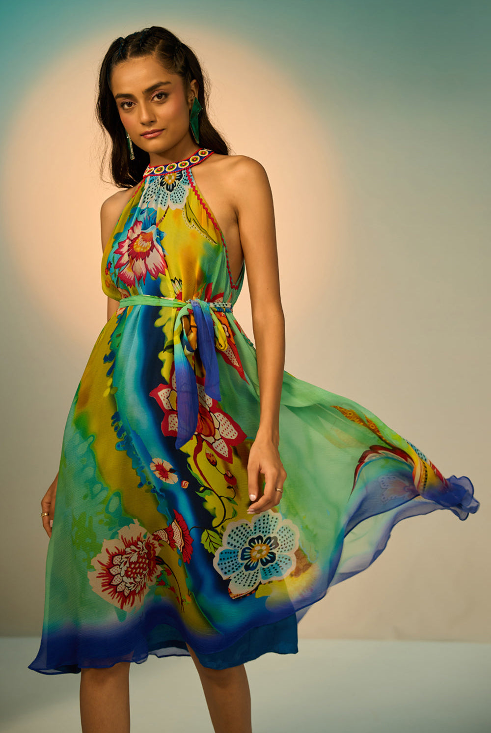 A woman modeling a sleeveless halter-neck midi dress with a vibrant multicolor floral and abstract print in shades of blue, green, yellow, and pink, styled with lime green pointed-toe heels against a studio backdrop.