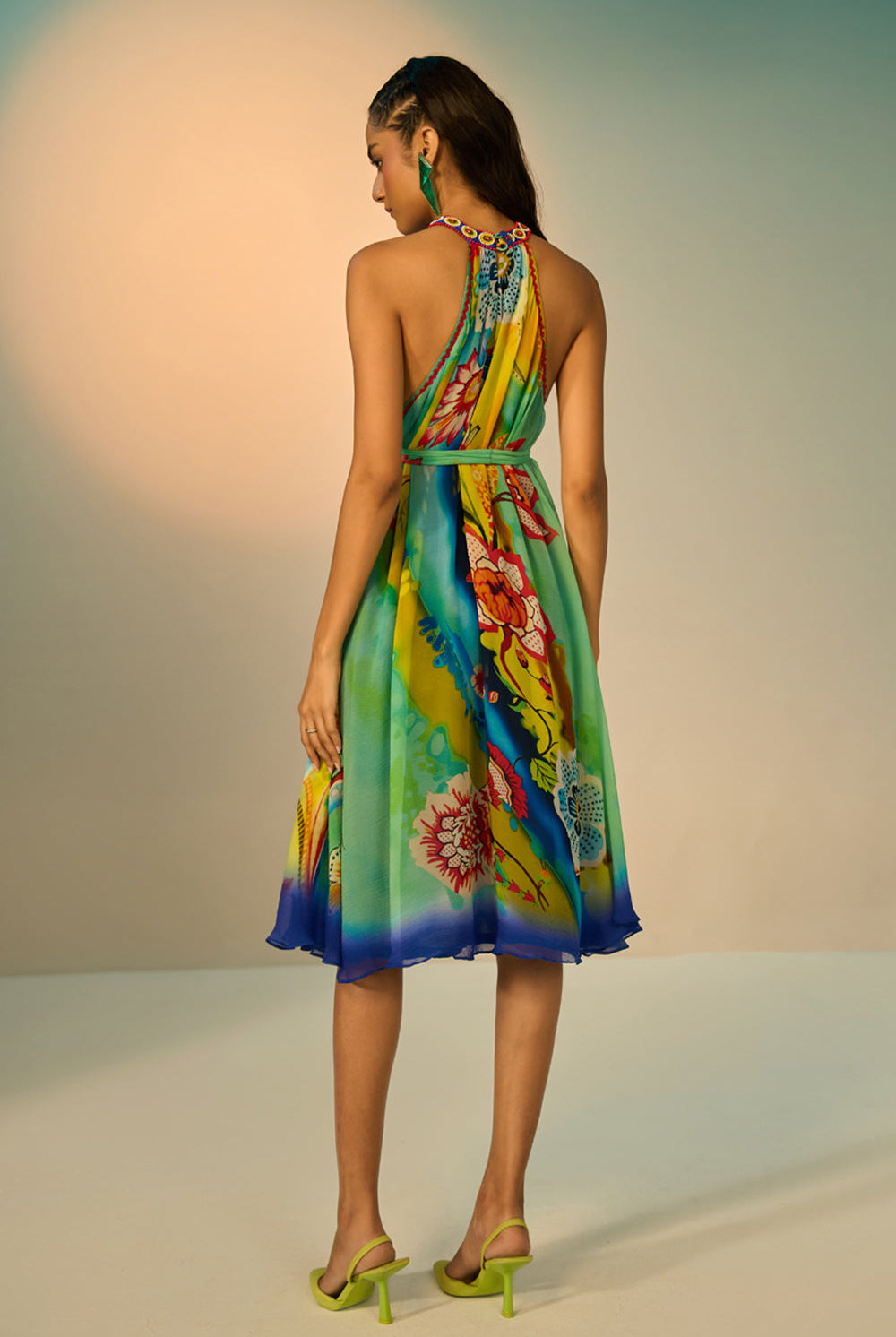 Back view of a woman modeling a sleeveless halter-neck midi dress with a vibrant multicolor floral and abstract print in shades of blue, green, yellow, and pink, styled with lime green pointed-toe heels against a studio backdrop.