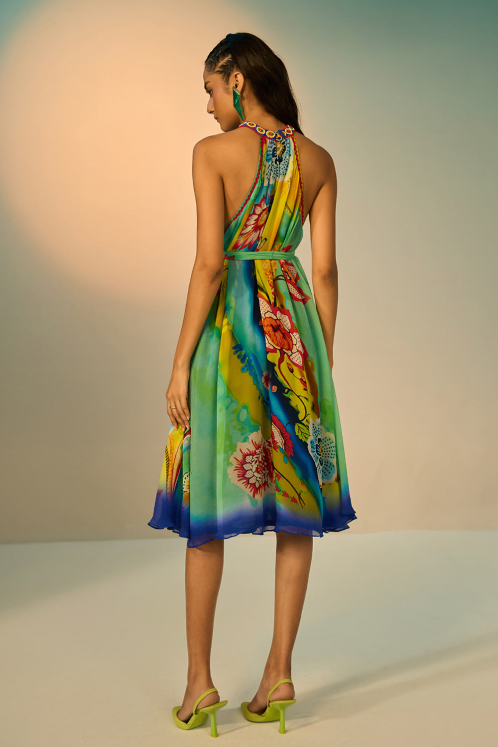 Back view of a woman modeling a sleeveless halter-neck midi dress with a vibrant multicolor floral and abstract print in shades of blue, green, yellow, and pink, styled with lime green pointed-toe heels against a studio backdrop.