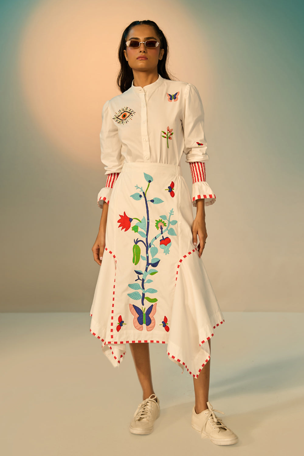 Woman wearing a ivory embroidered shirt with a skirt with colorful designs against a neutral background