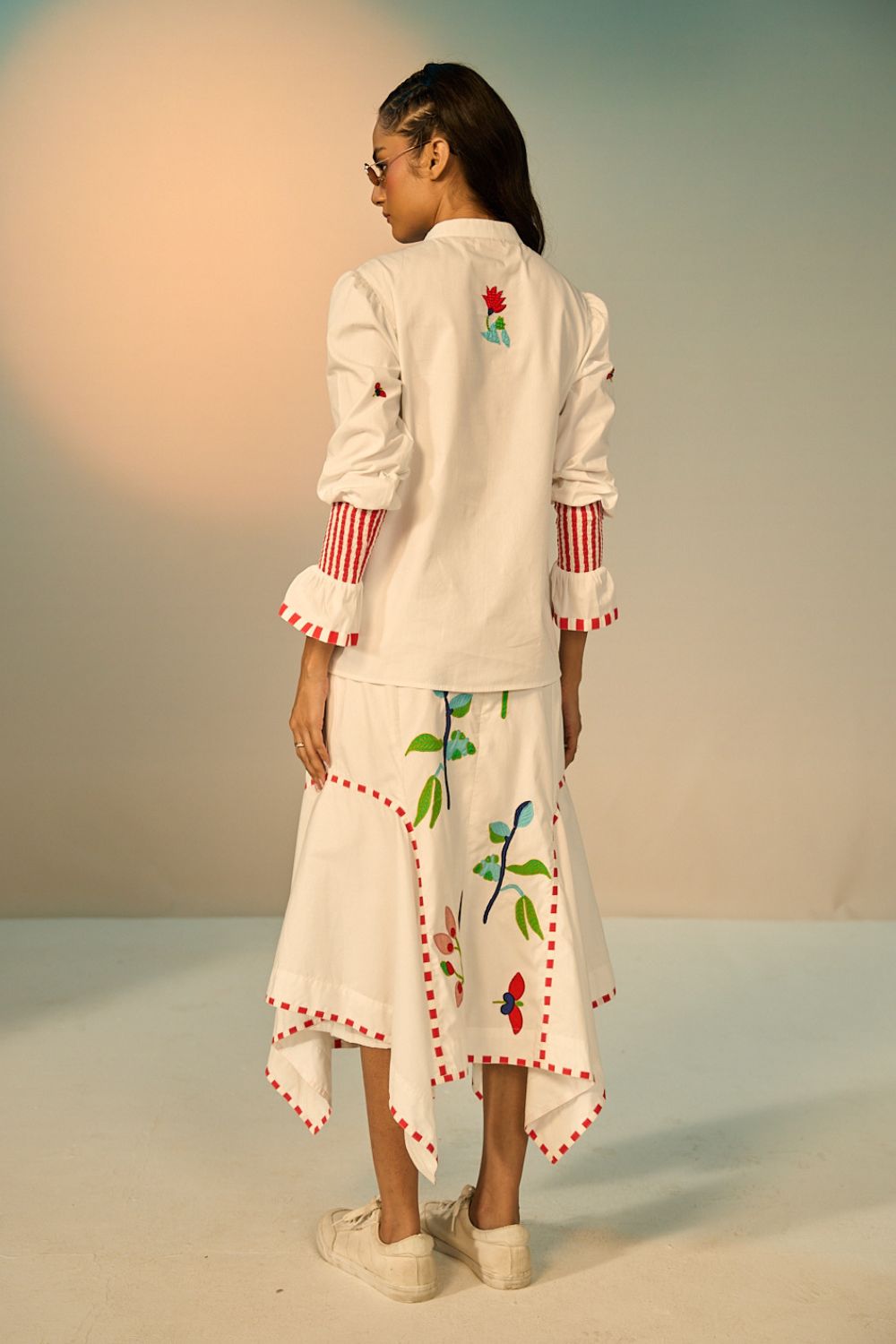 back view of a woman wearing a ivory embroidered shirt with a skirt with colorful designs against a neutral background