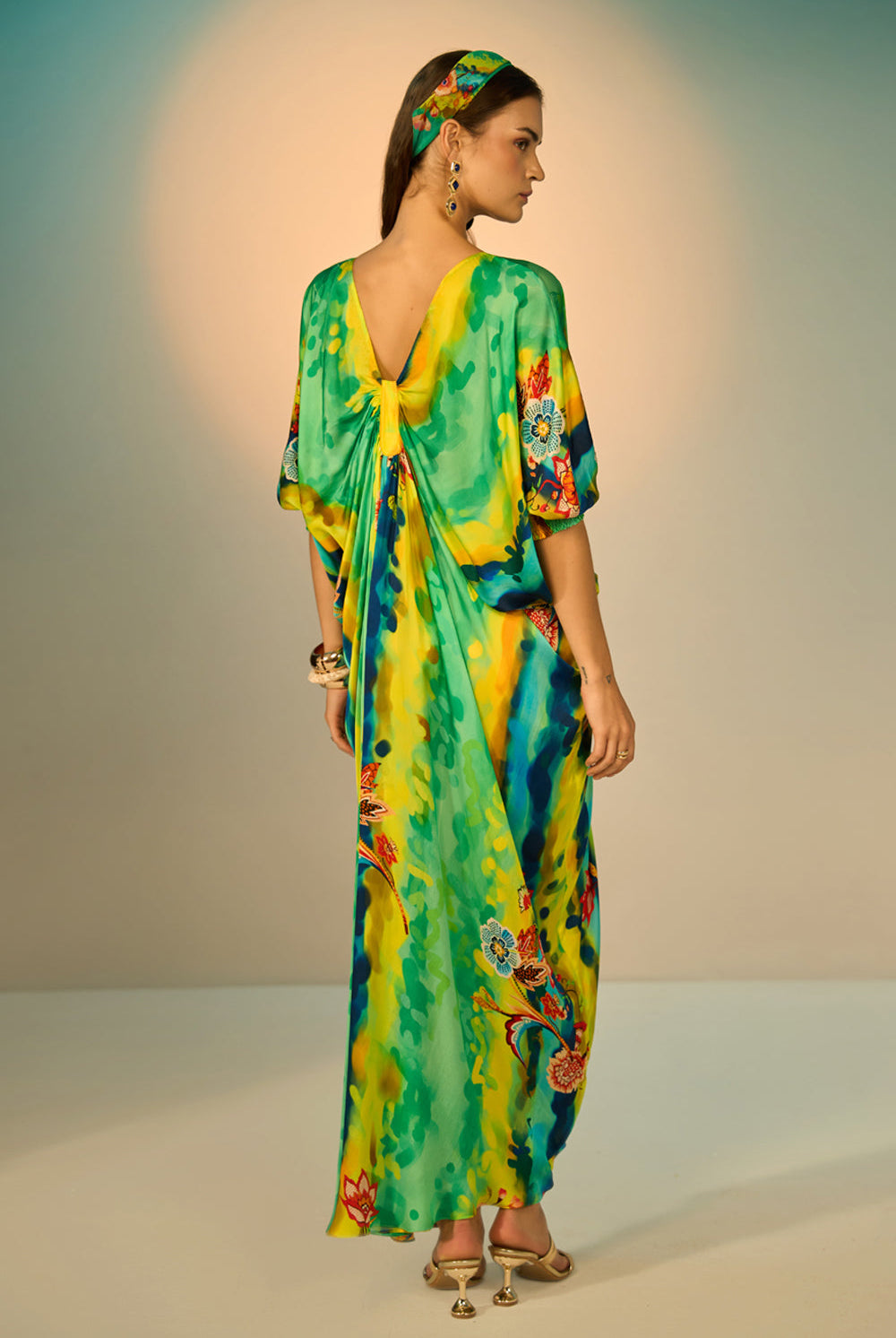 Back view of a woman modeling a vibrant, multicolored kaftan dress with a tie-dye pattern in multicolor, styled with a matching headband and gold accessories against a neutral studio backdrop.
