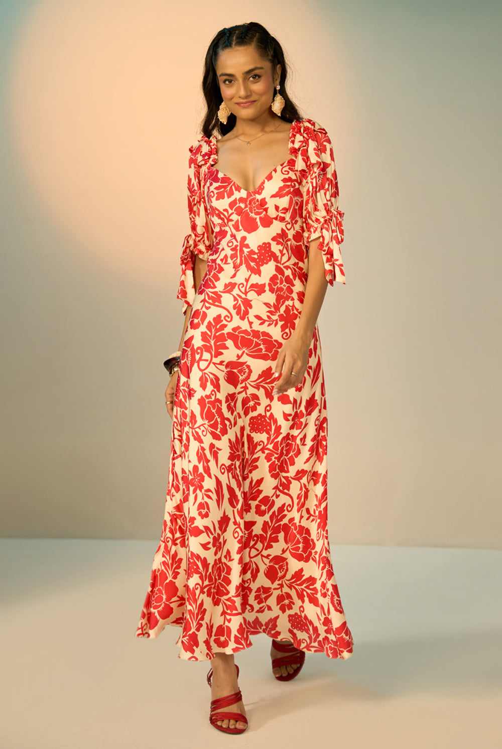 A woman modeling a red and cream floral print maxi dress with puff sleeves and a sweetheart neckline, standing against a neutral studio backdrop.