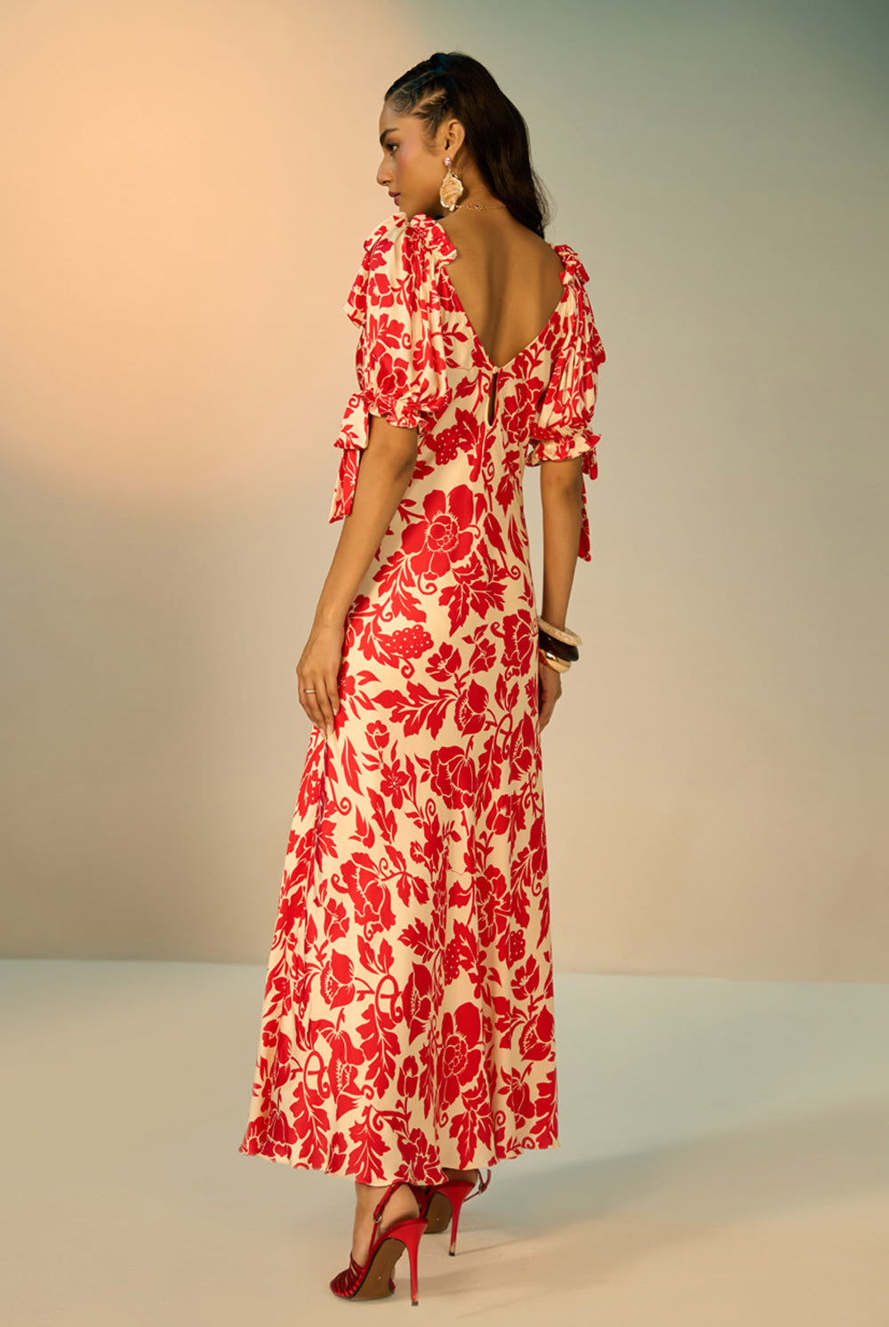 A woman modeling a red and cream floral print maxi dress with puff sleeves and a sweetheart neckline, standing against a neutral studio backdrop.