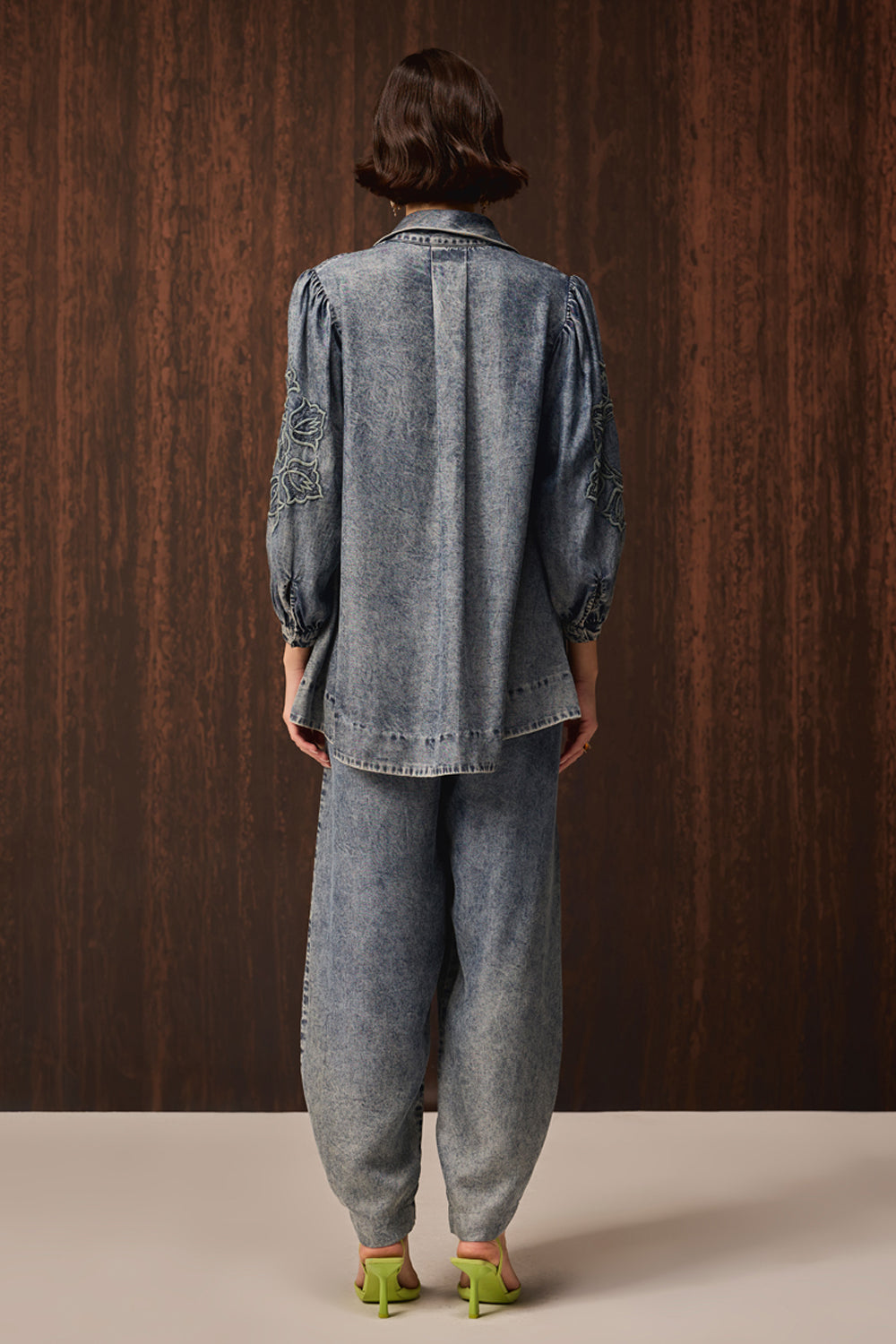 A model viewed from behind wearing a light blue denim two-piece set consisting of a long-sleeve button-up shirt with embroidered details on the sleeves and matching wide-leg trousers, paired with lime green heels against a dark wood-paneled background.
