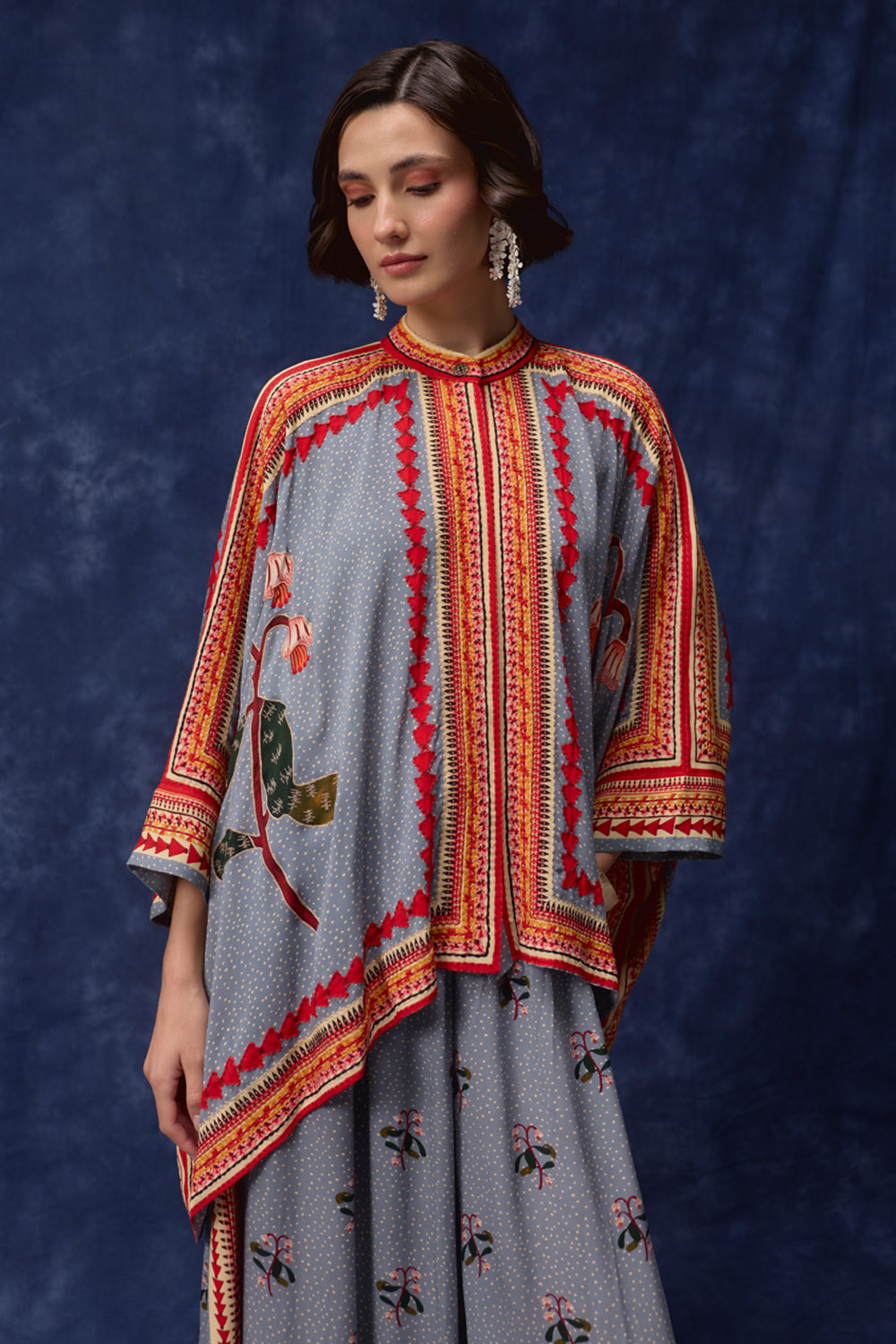 A woman models a traditional three-piece salwar kameez set featuring a gray base with red, orange, and gold geometric borders and floral embroidery, set against a dark blue textured backdrop.
