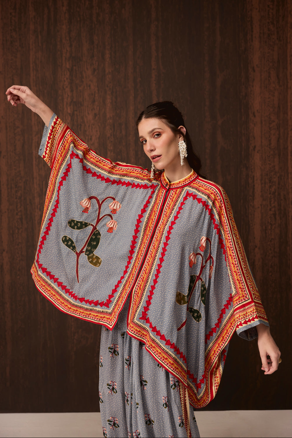 A woman models a gray patterned kaftan dress with wide sleeves, decorated with red and orange geometric borders and embroidered floral motifs, set against a dark wood-paneled background.