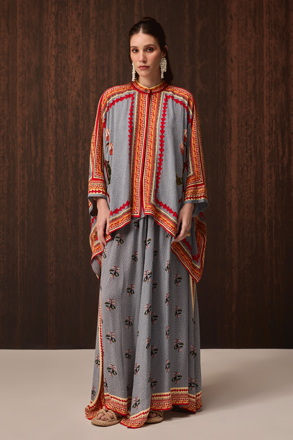 A woman models a traditional two-piece gray kurta set with orange and red embroidered borders and small black floral prints, set against a dark wood-paneled background.