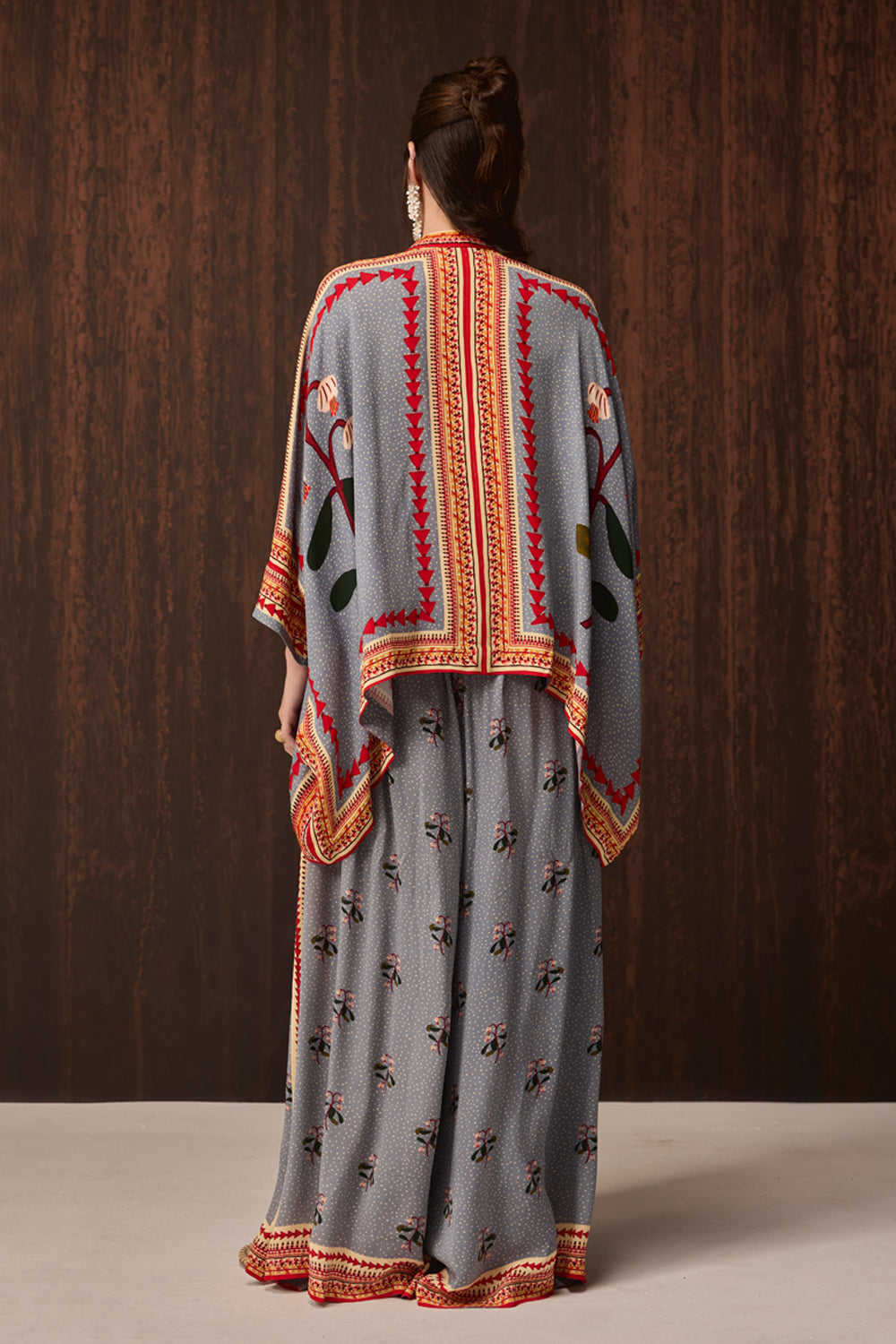 A woman viewed from behind wearing a gray traditional outfit with orange and red embroidered borders and floral motifs, standing against a dark wood-paneled background.