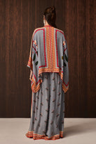 A woman viewed from behind wearing a gray traditional outfit with orange and red embroidered borders and floral motifs, standing against a dark wood-paneled background.