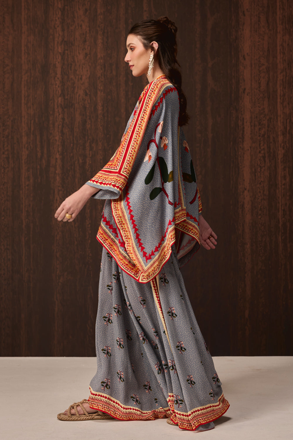 A woman models a traditional gray three-piece outfit with colorful embroidered borders and motifs, consisting of a kurta, flared pants, and a matching dupatta, set against a dark wood-paneled background.