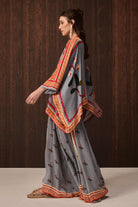 A woman models a traditional gray three-piece outfit with colorful embroidered borders and motifs, consisting of a kurta, flared pants, and a matching dupatta, set against a dark wood-paneled background.