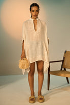 Woman wearing a beige linen dress holding a straw bag indoors.