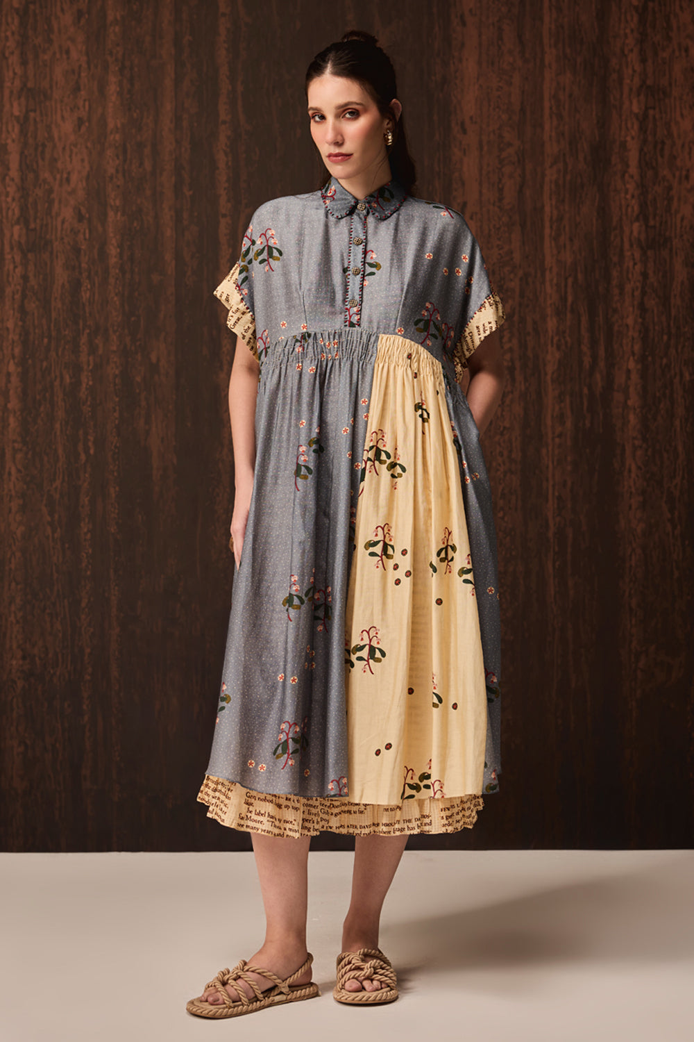 A woman modeling a short-sleeved midi dress with a smocked waist and a two-tone design of slate blue and cream with floral embroidery, standing against a dark wood-paneled background.