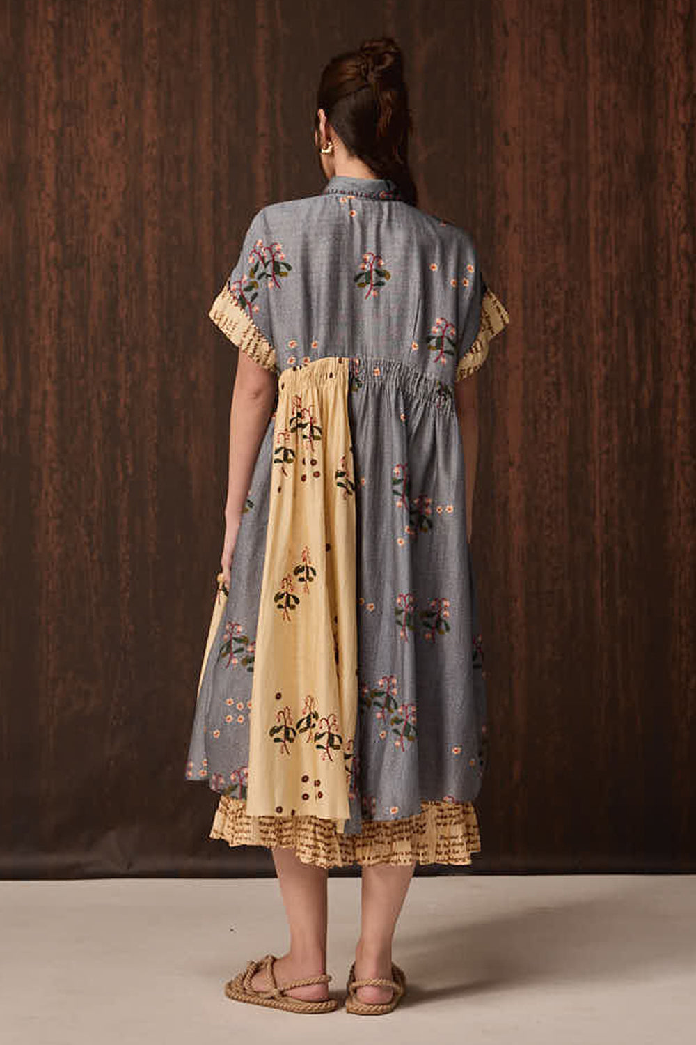 A woman viewed from behind wearing a short-sleeved midi dress with a blue-gray and beige color-blocked design and floral embroidery, standing against a dark wood-paneled background.