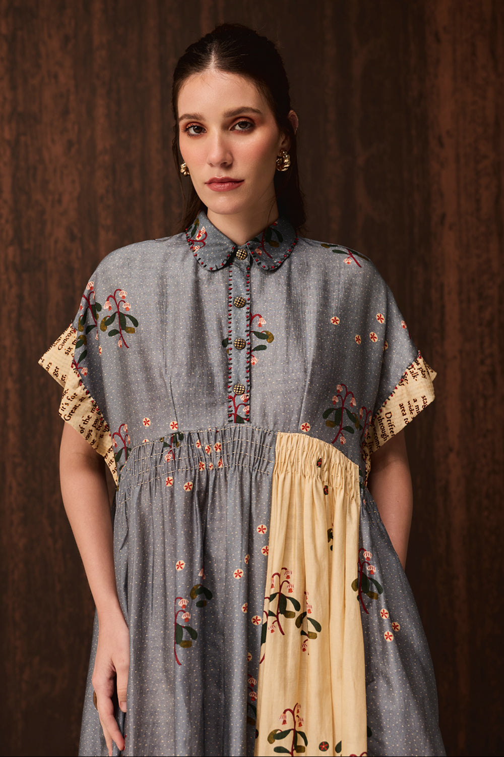 A woman wearing a short-sleeved dress with a gray floral upper section and a beige lower panel, posed against a dark wood background.