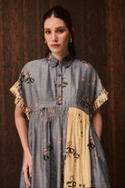A woman wearing a short-sleeved dress with a gray floral upper section and a beige lower panel, posed against a dark wood background.