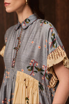 A close-up of a woman wearing a bohemian-style dress with a blue-gray chambray body, featuring floral embroidery, a beige ruffled collar, and a beige gathered waistband.