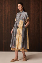 A woman models a short-sleeved midi dress with a blue-gray and yellow color-blocked design, featuring floral embroidery and lace trim, set against a dark wood-paneled background.