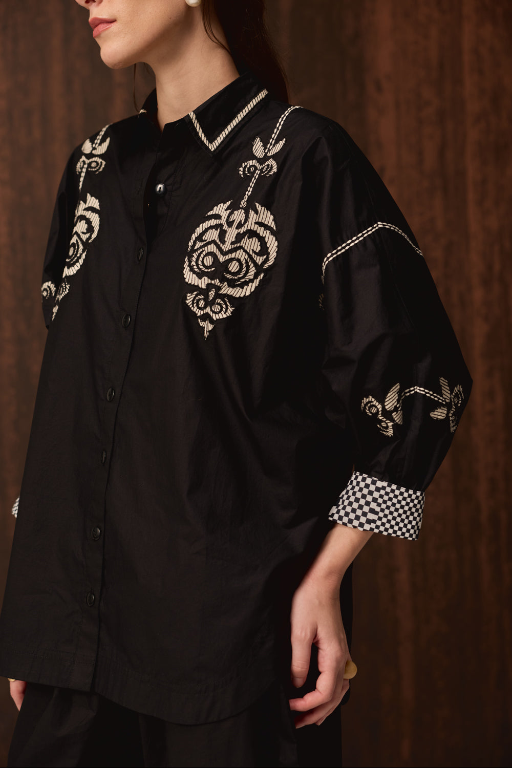 A close-up of a model wearing a black satin button-up shirt with intricate white embroidery on the chest and shoulders, a checkered cuff, and a pointed collar, set against a dark wood background.